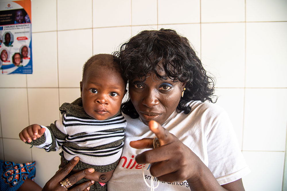 Dr. Nina Capo-Chichi holding a cleft-affected patient and pointing at the camera