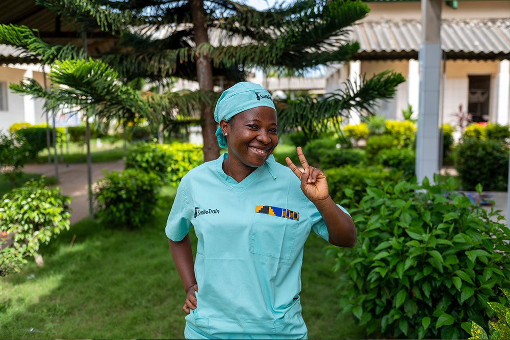 alt="Dr. Sanni standing outside in Smile Train scrubs smiling and flashing a peace sign"